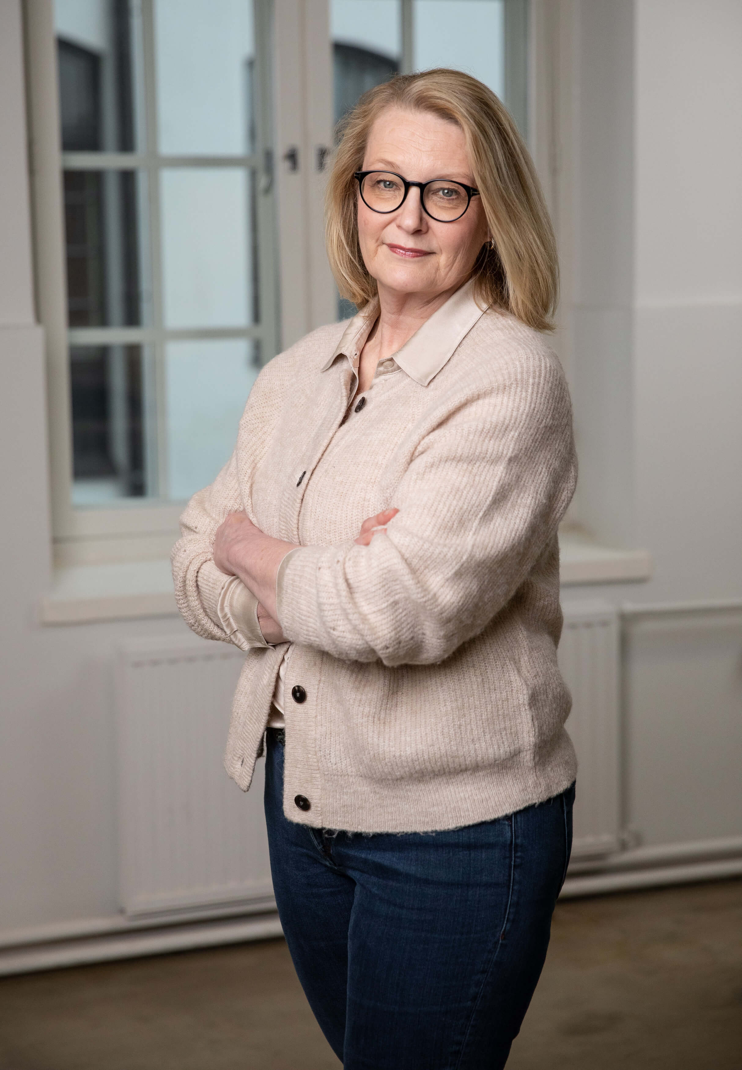 Nainen, jolla on vaaleat hiukset ja silmälasit, seisoo vasten valkoista seinää ja ikkunaa, kädet ristissä, pukeutuneena vaaleaan neuletakkiin ja farkkuihin.